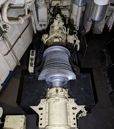 A large mechanical turbine with numerous metal blades is centrally positioned in a confined, industrial setting. Surrounding pipes, valves, and machinery suggest a technical or engineering environment, possibly within a power plant or ship engine room. The turbine features intricate details and components for mechanical operations.