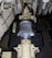 A large mechanical turbine with numerous metal blades is centrally positioned in a confined, industrial setting. Surrounding pipes, valves, and machinery suggest a technical or engineering environment, possibly within a power plant or ship engine room. The turbine features intricate details and components for mechanical operations.