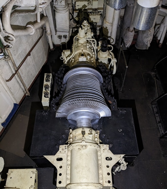 A large mechanical turbine with numerous metal blades is centrally positioned in a confined, industrial setting. Surrounding pipes, valves, and machinery suggest a technical or engineering environment, possibly within a power plant or ship engine room. The turbine features intricate details and components for mechanical operations.