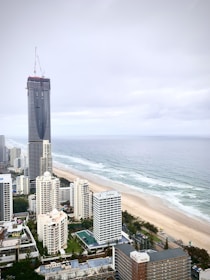 A coastal cityscape featuring a tall, under-construction skyscraper surrounded by other high-rise buildings near a long, sandy beach. The ocean meets the shoreline with gentle waves, and the sky is cloudy, indicating overcast weather.