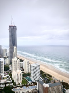 A coastal cityscape featuring a tall, under-construction skyscraper surrounded by other high-rise buildings near a long, sandy beach. The ocean meets the shoreline with gentle waves, and the sky is cloudy, indicating overcast weather.