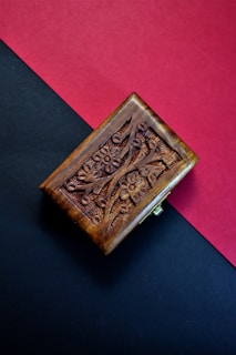 A wooden box with intricate floral carvings sits on a split background of red and black. The box is rectangular, with a rich brown hue and detailed craftsmanship evident in the ornate designs.