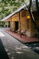 A rustic building with a thatched roof and beige-colored walls is situated in a lush, green environment. The path leading to the building is made of neatly arranged stone slabs. Several chairs and small tables are placed on a red concrete sitting area under the shelter of the building. Yellow hanging lights illuminate the area, and trees are integrated into the structure, adding to the natural aesthetic.