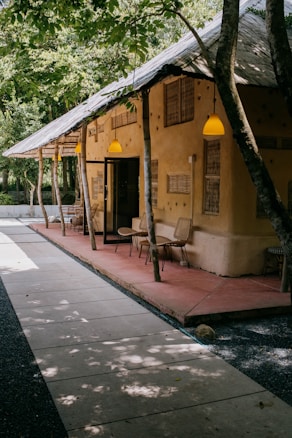 A rustic building with a thatched roof and beige-colored walls is situated in a lush, green environment. The path leading to the building is made of neatly arranged stone slabs. Several chairs and small tables are placed on a red concrete sitting area under the shelter of the building. Yellow hanging lights illuminate the area, and trees are integrated into the structure, adding to the natural aesthetic.