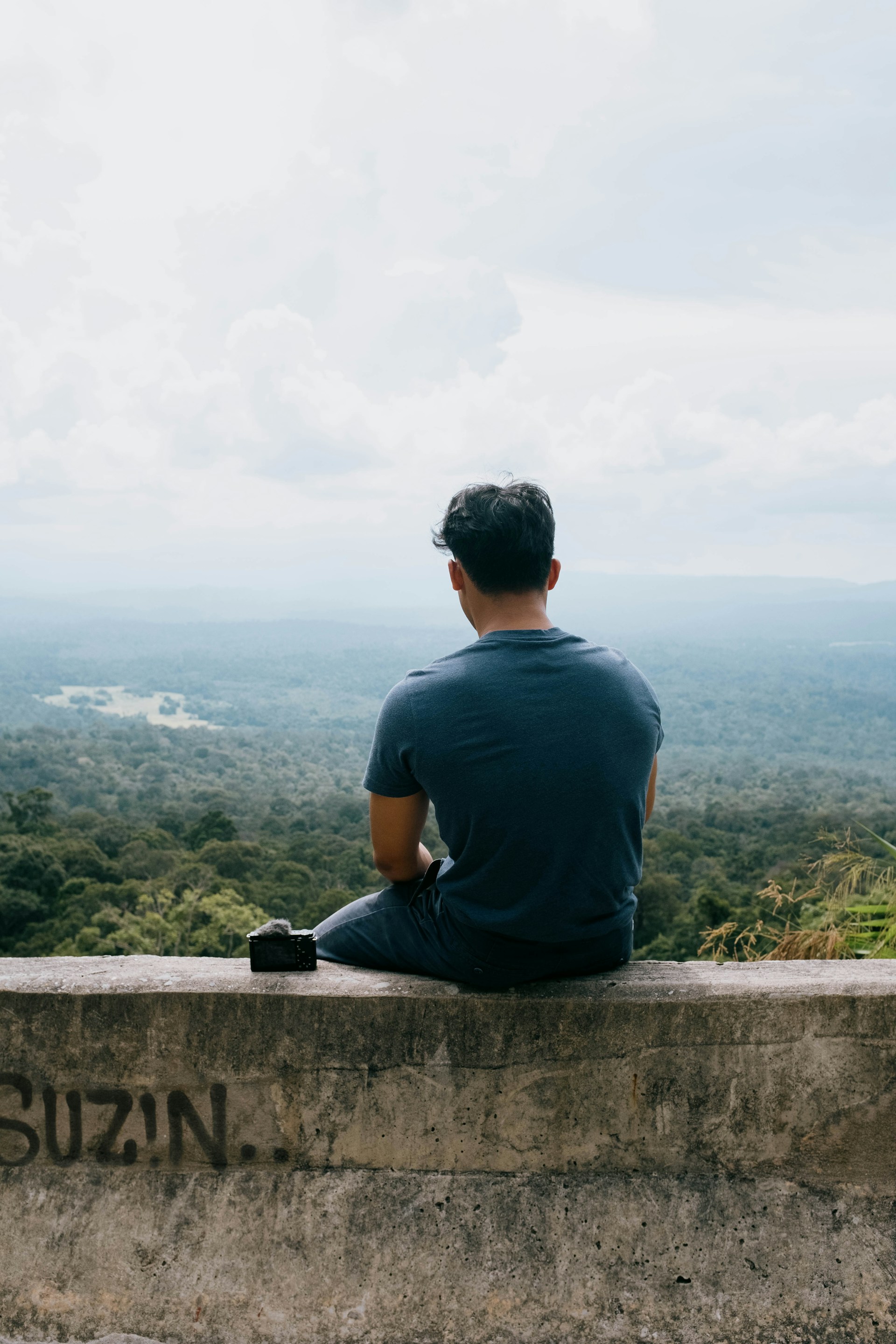 A person is seated on a concrete ledge, facing a vast panoramic view of lush green forests under a cloudy sky. A small black camera or accessory is placed next to the person on the ledge. The scene conveys a sense of tranquility and introspection.