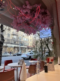 A chic cafe interior with red chairs and white tables is visible, adorned with cherry blossom decorations on the ceiling. The cafe has a modern vibe with elements like a 'MADE IN CHINA' neon sign seen through the window. Outside, a busy street features cars parked along the sidewalk and pedestrians walking.