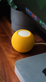 A yellow spherical smart speaker sits on a wooden desk. Next to it is a sleek, silver computer monitor base. In the background, a gray planter is partially visible.