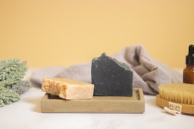 Two bars of soap, one beige and one black, are displayed on a rectangular tray. Behind them is a gray cloth, an amber glass dropper bottle, a wooden brush with bristles, and a cluster of green foliage. The background is a soft yellow color.