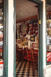 An interior of a vintage-themed diner is visible through an open door. The wall is adorned with retro Coca-Cola memorabilia and old posters. Wooden chairs are neatly lined up against a counter. The floor is covered with a black and white checkered pattern, enhancing the retro ambiance.