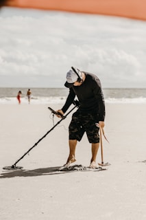 A person is using a metal detector on a sandy beach. The individual is wearing a black shirt, camo shorts, a cap, and headphones. In the background, the ocean and two other people walking are visible. The sky is partly cloudy.