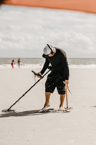 A person is using a metal detector on a sandy beach. The individual is wearing a black shirt, camo shorts, a cap, and headphones. In the background, the ocean and two other people walking are visible. The sky is partly cloudy.