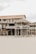 A rustic, wooden beach house elevated on stilts is set against the backdrop of an overcast sky. The house features a straw-thatched awning and a balcony with lattice railings. Its weathered appearance and natural surroundings suggest a serene, seaside location.