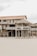 A rustic, wooden beach house elevated on stilts is set against the backdrop of an overcast sky. The house features a straw-thatched awning and a balcony with lattice railings. Its weathered appearance and natural surroundings suggest a serene, seaside location.