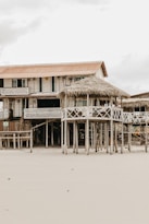 A rustic, wooden beach house elevated on stilts is set against the backdrop of an overcast sky. The house features a straw-thatched awning and a balcony with lattice railings. Its weathered appearance and natural surroundings suggest a serene, seaside location.