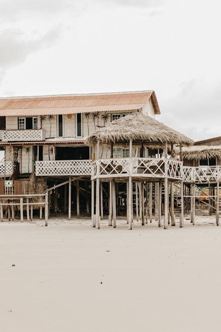 A rustic, wooden beach house elevated on stilts is set against the backdrop of an overcast sky. The house features a straw-thatched awning and a balcony with lattice railings. Its weathered appearance and natural surroundings suggest a serene, seaside location.