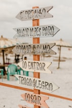 A wooden sign with multiple directional arrows featuring inspirational Portuguese phrases is placed on a sandy beach. In the background, there are beach umbrellas and several plastic chairs, creating a relaxed and inviting atmosphere.