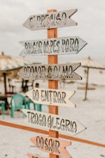 A wooden sign with multiple directional arrows featuring inspirational Portuguese phrases is placed on a sandy beach. In the background, there are beach umbrellas and several plastic chairs, creating a relaxed and inviting atmosphere.