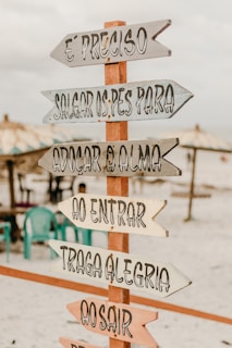 A wooden sign with multiple directional arrows featuring inspirational Portuguese phrases is placed on a sandy beach. In the background, there are beach umbrellas and several plastic chairs, creating a relaxed and inviting atmosphere.