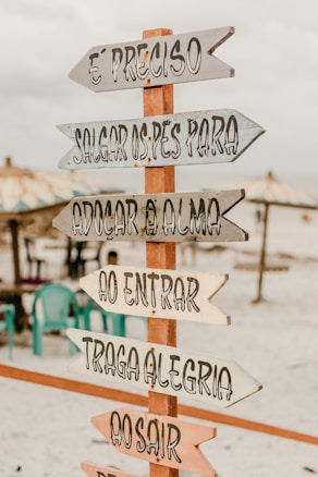 A wooden sign with multiple directional arrows featuring inspirational Portuguese phrases is placed on a sandy beach. In the background, there are beach umbrellas and several plastic chairs, creating a relaxed and inviting atmosphere.