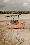 A small, vibrant orange wooden boat is resting on a sandy shore. The boat has blue accents and a weathered appearance, with a simple canopy structure on top. The words 'Vou Com Deus' are painted on the side. Sparse vegetation grows in the sand, and a dense line of trees is visible in the background under a cloudy sky.