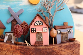 A miniature scene with tiny wooden houses painted in soft pastel colors and small models of trees and a windmill. There is a pink house with a brown roof, a blue house, and a small white house. The background features a bright blue sky with the sun partially visible. The trees are crafted from dried materials, adding texture to the composition.