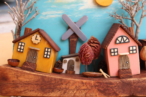 A whimsical scene features miniature houses crafted from wood with charming details such as windows and doors. One house is painted yellow with a small clock, and another is pink. Surrounding the houses are tree-like structures, and the background depicts a blue sky with a sun and clouds.