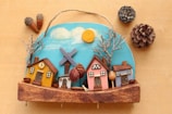 A handcrafted wooden decoration featuring a small village scene with colorful houses, bare trees, and a windmill against a painted blue sky with clouds and a bright sun. Pinecones and acorns are arranged around the decoration, adding a natural element to the scene.