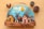 A handcrafted wooden decoration featuring a small village scene with colorful houses, bare trees, and a windmill against a painted blue sky with clouds and a bright sun. Pinecones and acorns are arranged around the decoration, adding a natural element to the scene.