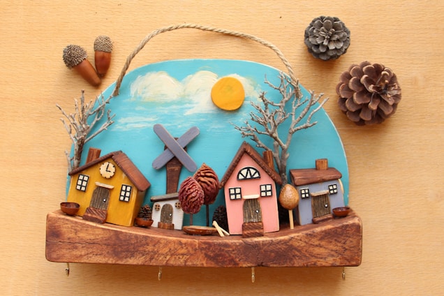A handcrafted wooden decoration featuring a small village scene with colorful houses, bare trees, and a windmill against a painted blue sky with clouds and a bright sun. Pinecones and acorns are arranged around the decoration, adding a natural element to the scene.