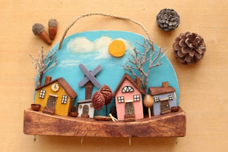 A handcrafted wooden decoration featuring a small village scene with colorful houses, bare trees, and a windmill against a painted blue sky with clouds and a bright sun. Pinecones and acorns are arranged around the decoration, adding a natural element to the scene.