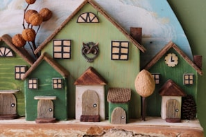 A charming, handcrafted wooden village scene with several small houses painted in green and beige tones. Each house has distinct features such as wooden doors, small windows, and a roof with a natural, rustic texture. An owl decoration adds a whimsical touch, and dried seed pods resemble trees or foliage. A painted clock shows a time of approximately 1 o'clock.