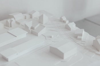 A minimalist architectural model featuring a small urban layout with simplified building forms. The model is monochromatic, focusing on structure and spatial arrangement. The image emphasizes clean lines and a serene aesthetic.