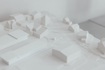 A minimalist architectural model featuring a small urban layout with simplified building forms. The model is monochromatic, focusing on structure and spatial arrangement. The image emphasizes clean lines and a serene aesthetic.