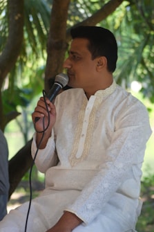 A person wearing a traditional white kurta is holding a microphone while seated outdoors under trees. The background is lush with green foliage, suggesting a natural, serene setting.