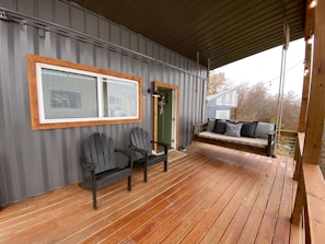 A cozy porch with wooden flooring and a metal siding wall. Two black wooden chairs are positioned next to a wide window with a wooden frame. A hanging daybed with gray cushions is suspended by ropes. String lights are hanging above, adding a warm atmosphere. The porch has a view of autumn trees and another building in the background.