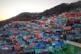 A vibrant hillside village featuring an array of colorful houses tightly packed together. The houses are painted in vivid hues such as blue, pink, green, and yellow, creating a striking visual effect. The backdrop includes forested hills under a pale sky with hints of sunset light.