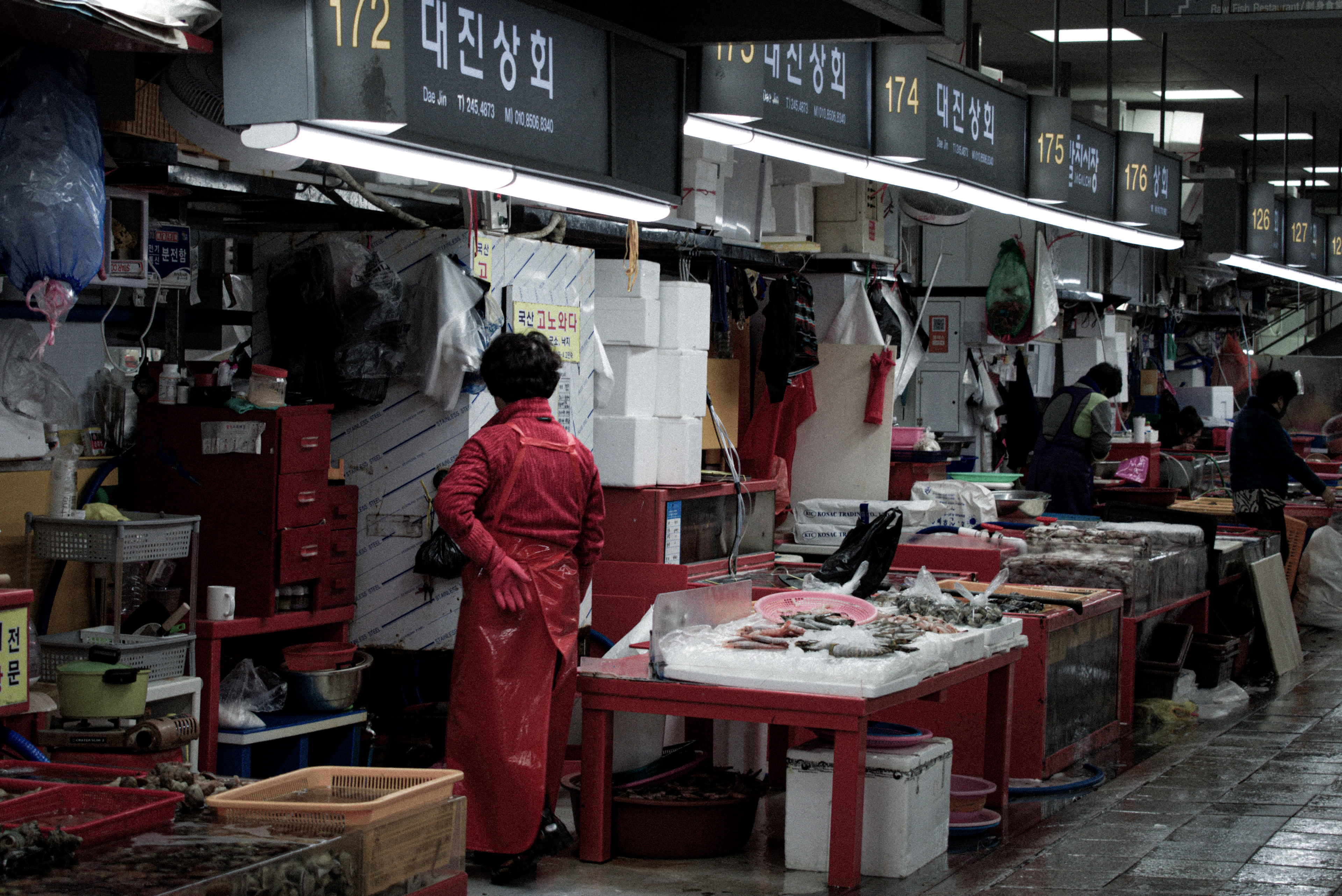 A fishmonger in red waders among the stalls of Jagalchi Fish Market in Busan, trays of fresh fish laid out along the aisle