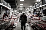 An indoor fish market scene with a person wearing a helmet and waterproof overalls walking down an aisle. Various fish and seafood are displayed on both sides in tanks and on tables. The market has fluorescent lighting and visible signage with numbers and text in an Asian script.