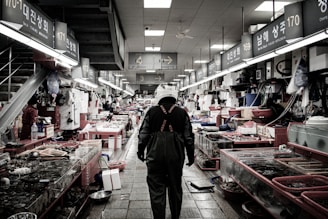 An indoor fish market scene with a person wearing a helmet and waterproof overalls walking down an aisle. Various fish and seafood are displayed on both sides in tanks and on tables. The market has fluorescent lighting and visible signage with numbers and text in an Asian script.