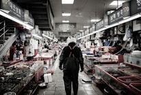 An indoor fish market scene with a person wearing a helmet and waterproof overalls walking down an aisle. Various fish and seafood are displayed on both sides in tanks and on tables. The market has fluorescent lighting and visible signage with numbers and text in an Asian script.