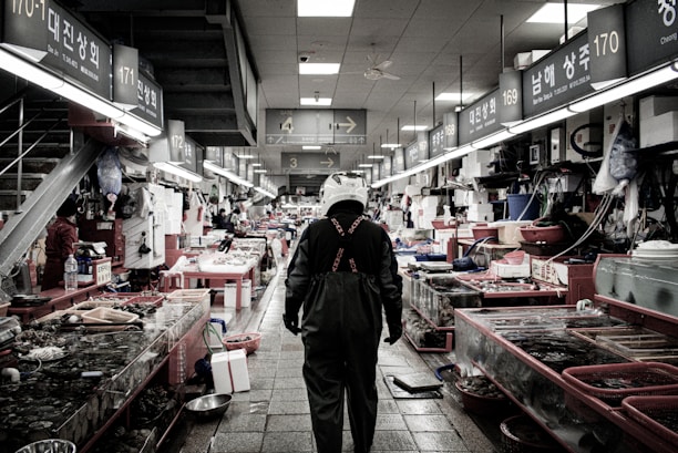 An indoor fish market scene with a person wearing a helmet and waterproof overalls walking down an aisle. Various fish and seafood are displayed on both sides in tanks and on tables. The market has fluorescent lighting and visible signage with numbers and text in an Asian script.