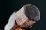 A close-up image of the head of a well-used hammer with a rustic, weathered surface. The metal appears rough with visible rust and marks, set against a dark background, emphasizing the texture and ruggedness of the tool.