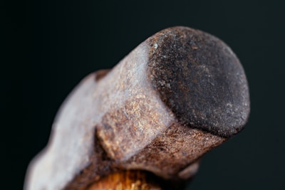 A close-up image of the head of a well-used hammer with a rustic, weathered surface. The metal appears rough with visible rust and marks, set against a dark background, emphasizing the texture and ruggedness of the tool.