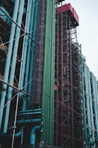 A complex structure with a series of colorful pipes and metal scaffolding. Various hues of blue, green, and red dominate the tall, vertical arrangement. The industrial elements create a maze-like pattern, with pipes crossing and intersecting amidst metallic frameworks. The setting suggests a part of an industrial facility or building dedicated to utility systems.
