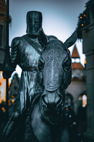 A knight wearing medieval armor is seated on a horse. The knight's helmet obscures his face, and intricate designs cover both the knight’s armor and the horse's tack. The setting appears to be a historical or fantasy-themed location, with blurred architectural elements visible in the background.