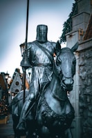 A statue of a knight in full armor sitting atop a horse, holding a lance. The knight's face is covered by a helmet, and intricate details are visible on both the knight's armor and the horse's bridle. The background includes a stone facade with decorative elements and what appears to be medieval-style architecture, along with faintly visible festive lights suggesting a time around the holidays.