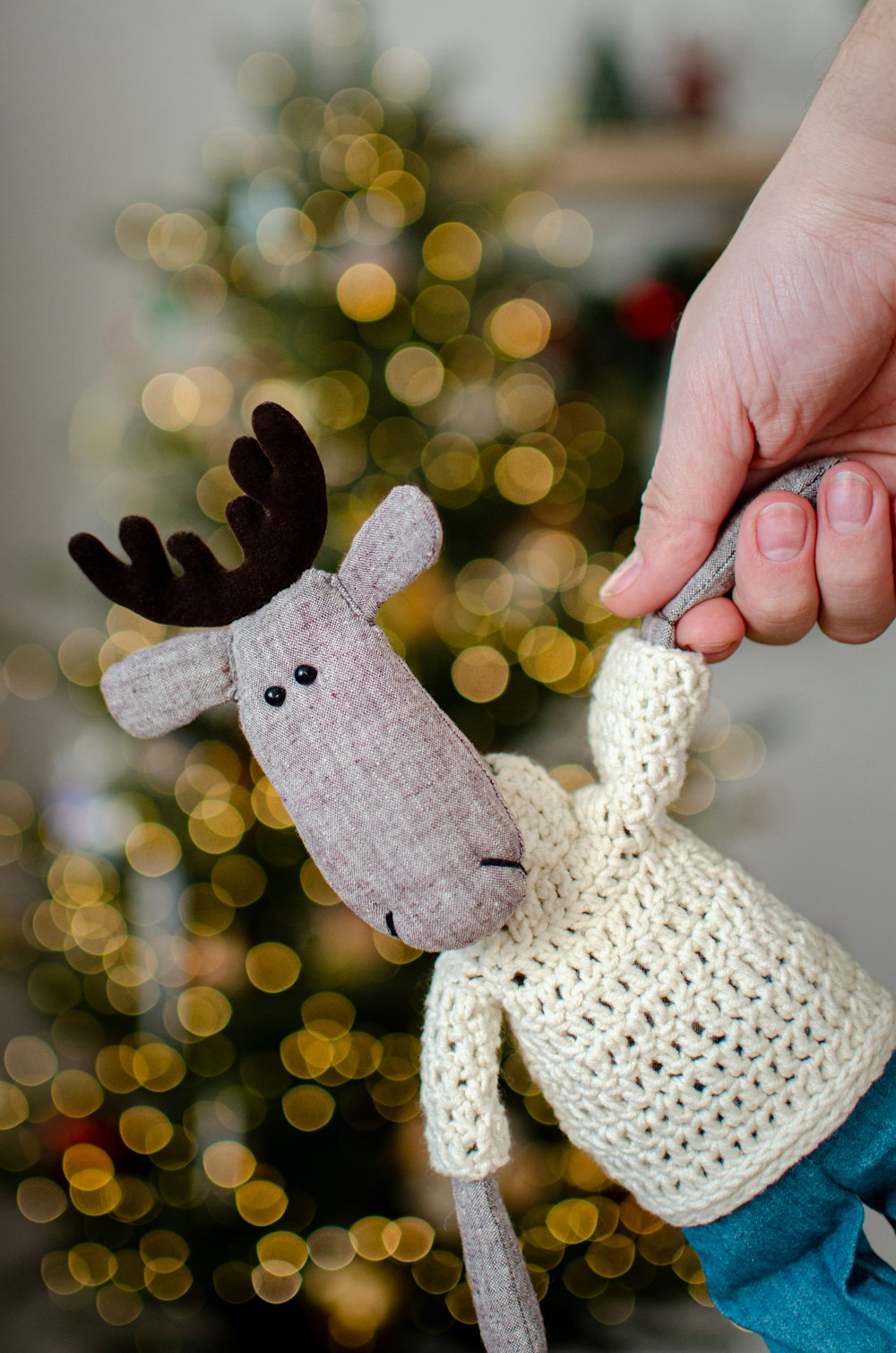a hand holding a knitted moose ornament in front of a christmas tree