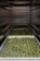 Several trays filled with green cannabis buds are placed on shelves within a metallic enclosure. The buds are arranged densely, and the perspective gives a view down the narrow row created by the shelves.