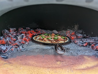 A cast-iron pan with a pizza topped with slices of green jalapeño, sausage, and tomato is placed inside a wood-fired oven. The flames and glowing embers from the burning wood create an intense heat, contrasting with the dark interior of the oven.