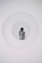 A person stands facing away, positioned centrally within a series of concentric circular frames giving a surreal, tunnel-like visual effect. The individual is wearing a plaid dress, contributing a vintage or classic tone to the minimalist and futuristic setting.